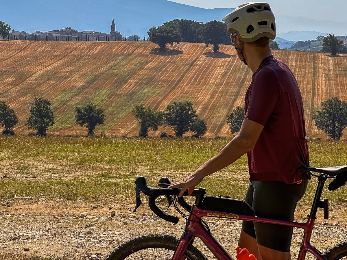 Un ciclista in piedi accanto alla bici osserva campi e colline umbre, con un borgo e il suo campanile all'orizzonte