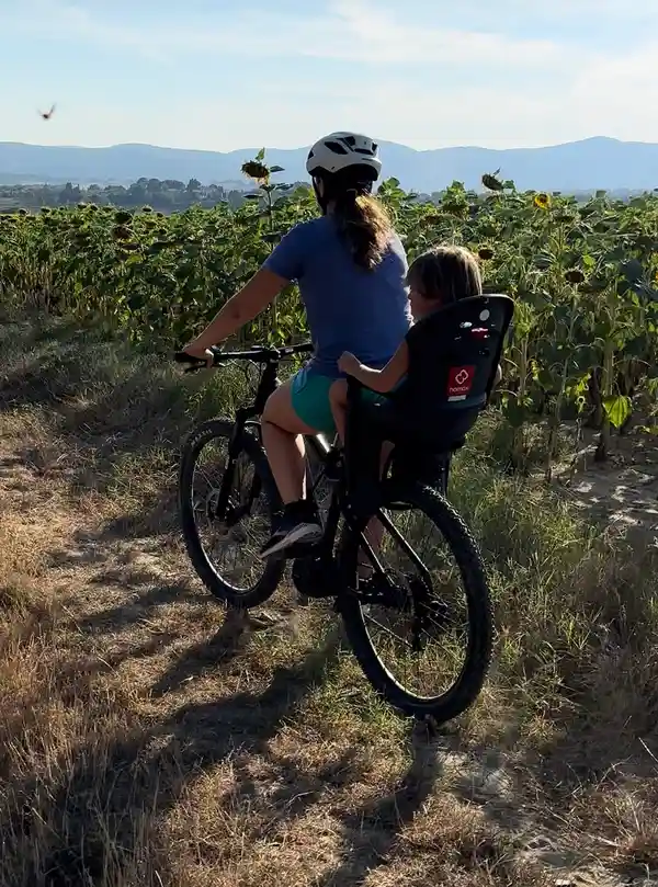Mamma in bici col casco e il bambino nel seggiolino, tra i girasoli